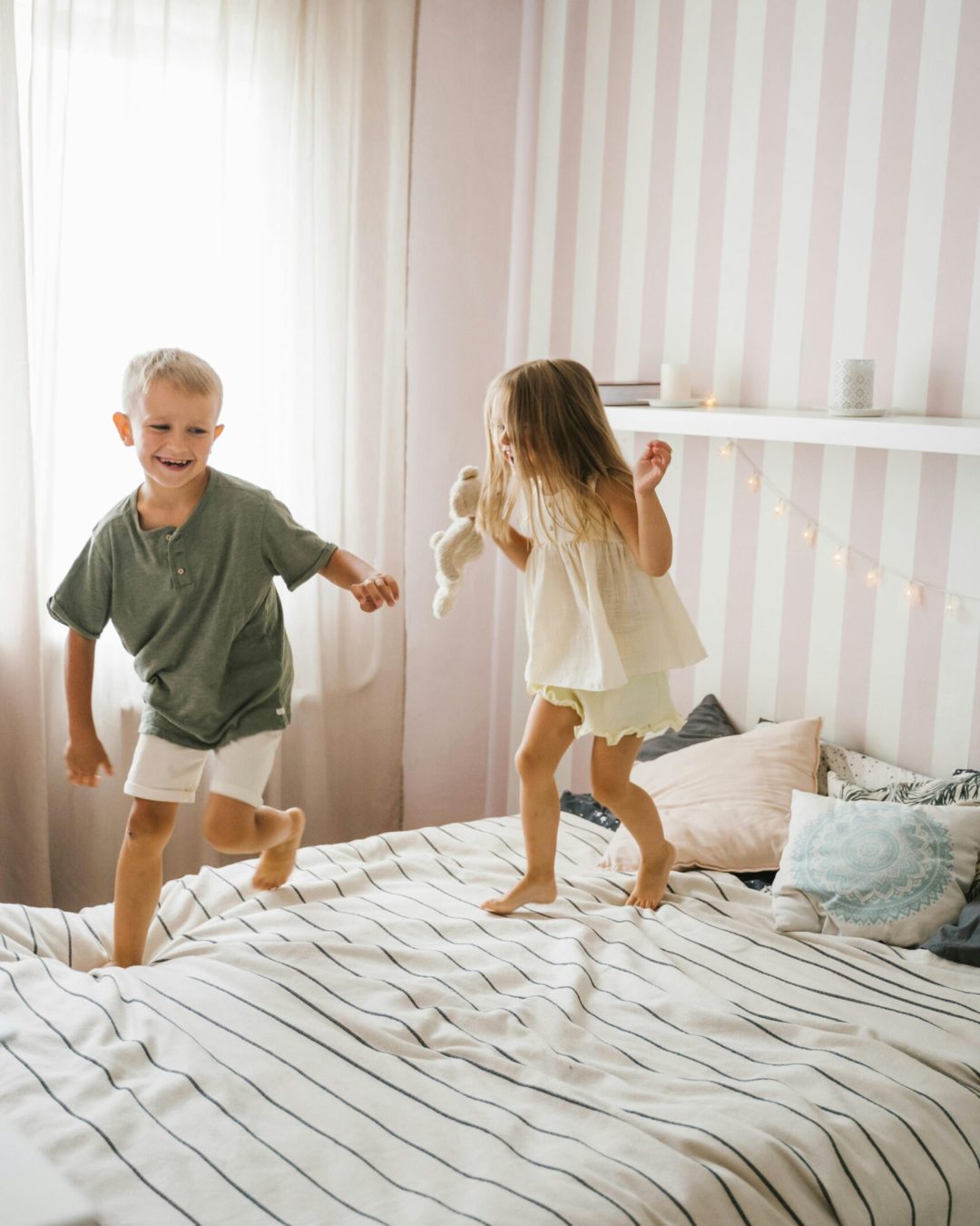 Two kids joyfully jumping on bed in a bright room with soft lighting and playful decor.